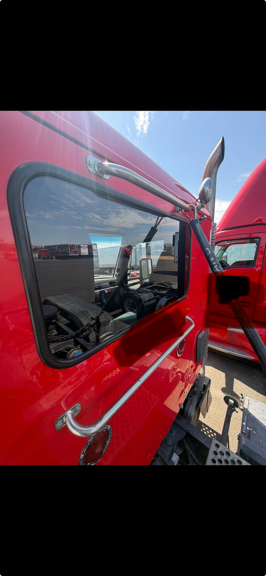 Close-up of a red semi-truck cab showing the side window, chrome grab handles, side mirror, and part of the front hood in an outdoor setting.